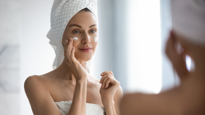 Attractive young adult woman applying facial cream looking in mirror