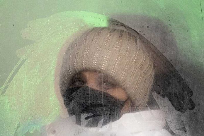 woman wearing beanie and face mask looking out a fogged up window