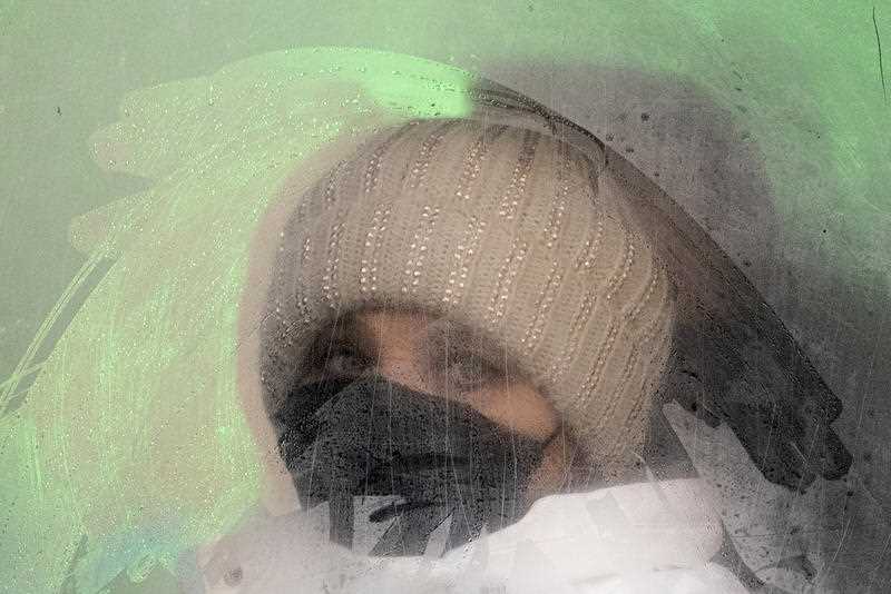 woman wearing beanie and face mask looking out a fogged up window